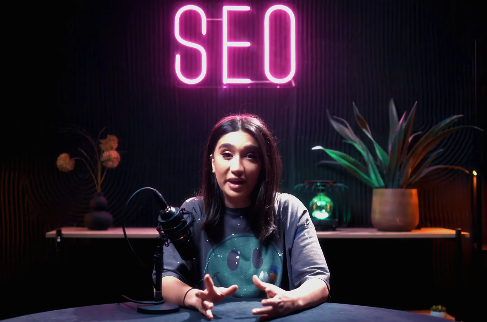 A woman speaking into a microphone in a dimly lit studio with a neon sign reading 'SEO' above her.